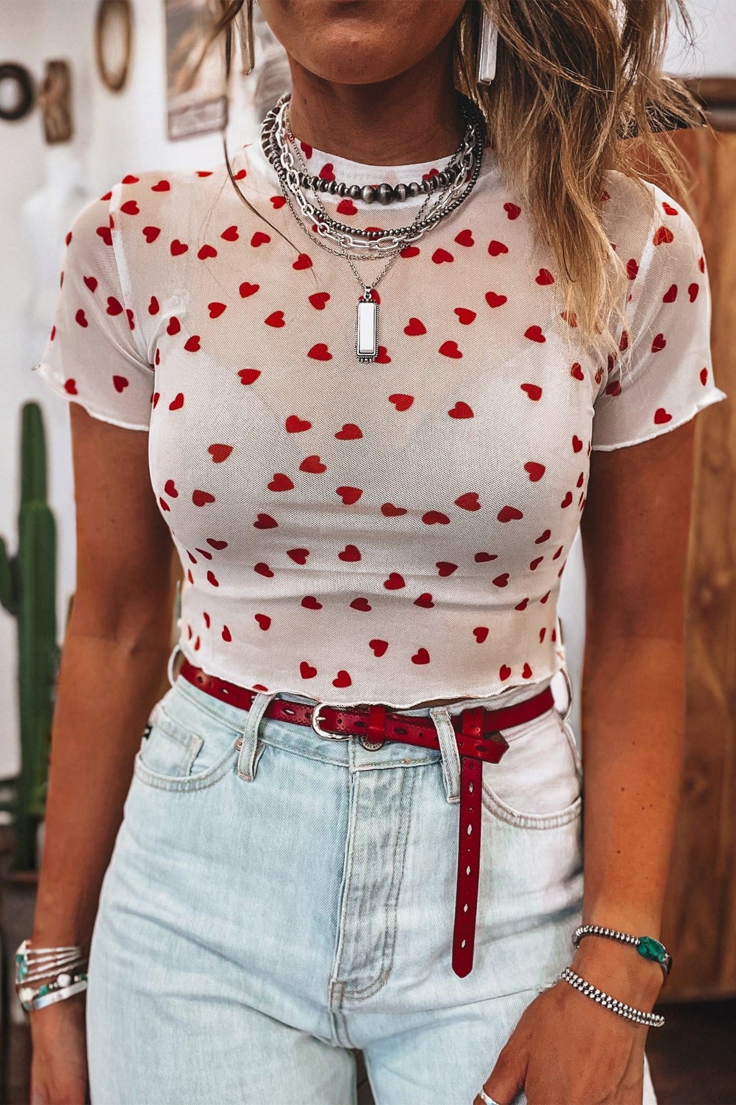 A stylish woman wearing a white mesh short sleeve top with an allover red heart print, layered under a denim vest.