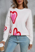 A woman wearing a white long sleeve sweater with a vibrant pink and red heart pattern, styled with light blue jeans.