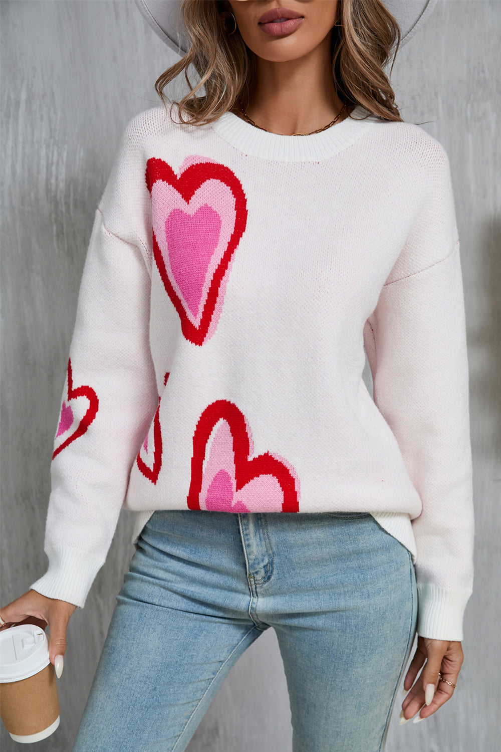 A woman wearing a white long sleeve sweater with a vibrant pink and red heart pattern, styled with light blue jeans.