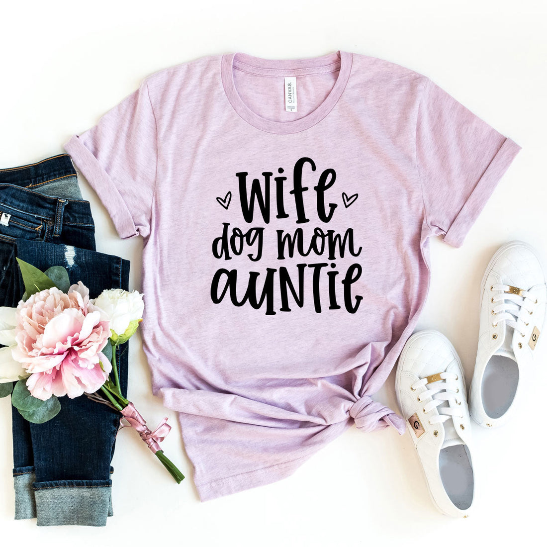 A heather mauve t-shirt with black text that reads Wife, Dog Mom, Auntie placed next to a bouquet of flowers and white sneakers.