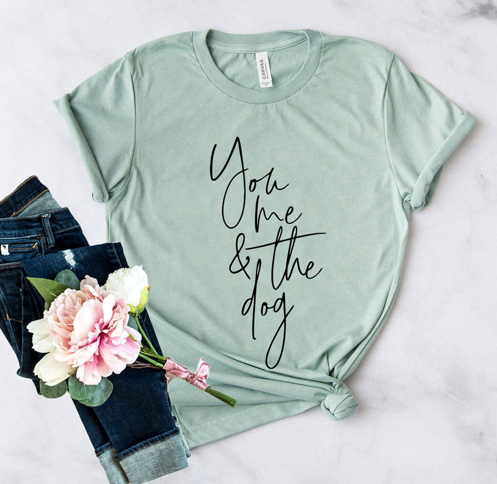 Sage green graphic t-shirt with the text You Me And The Dog in black script, displayed with jeans and flowers.