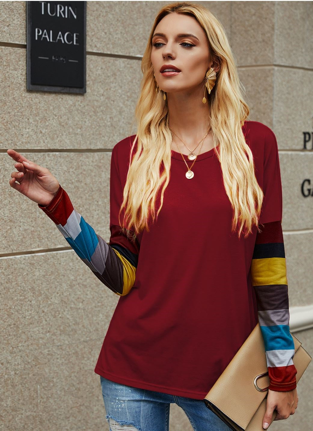 A woman wearing a maroon long sleeve sweater with multi-colored striped sleeves, paired with denim jeans.