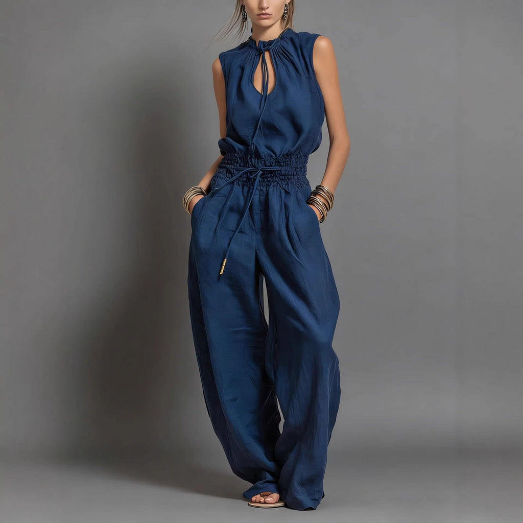 Model wearing a navy blue sleeveless high-waist wide-leg jumpsuit with a tie-waist detail on a city street.