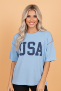 A smiling woman wearing a light blue corded knit short sleeve T-shirt with the word USA printed in dark blue on the front.