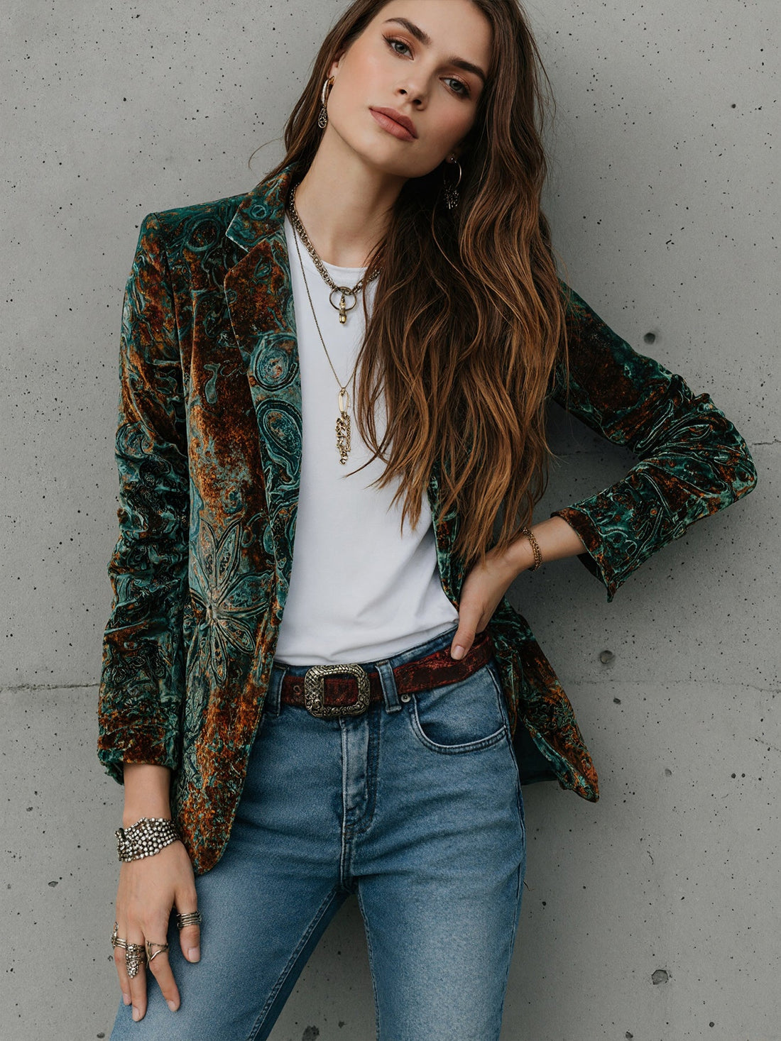 A woman wearing a stylish green and copper colored printed velvet blazer, layered over a white t-shirt and blue jeans.