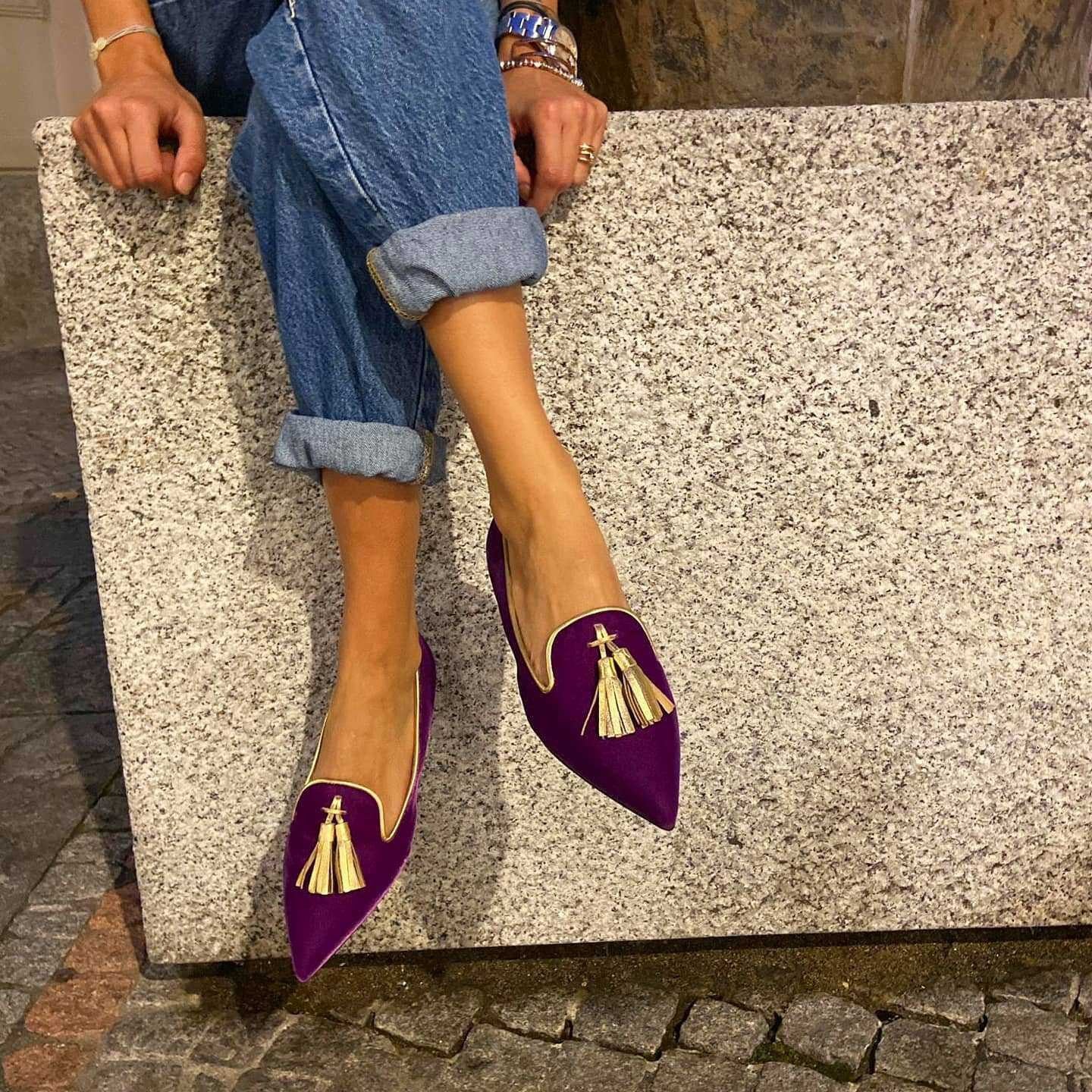 Close up of vibrant purple suede pointed toe flats featuring gold tassels, worn with cuffed blue jeans.