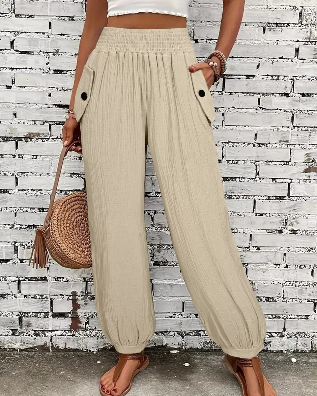 Person wearing beige wide-leg pants with a white top and brown sandals, holding a woven bag against a brick wall.