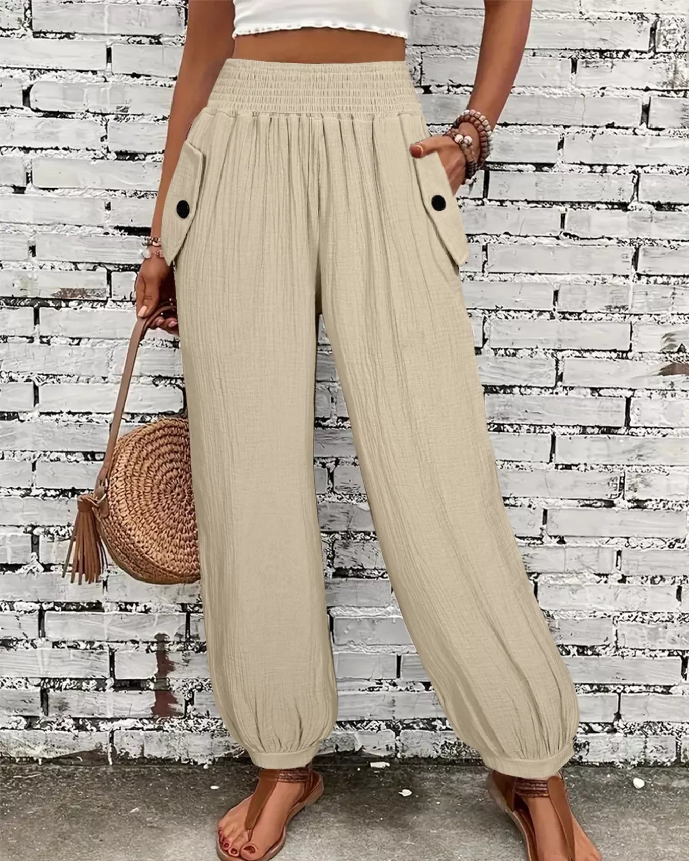 Person wearing beige wide-leg pants with a white top and brown sandals, holding a woven bag against a brick wall.