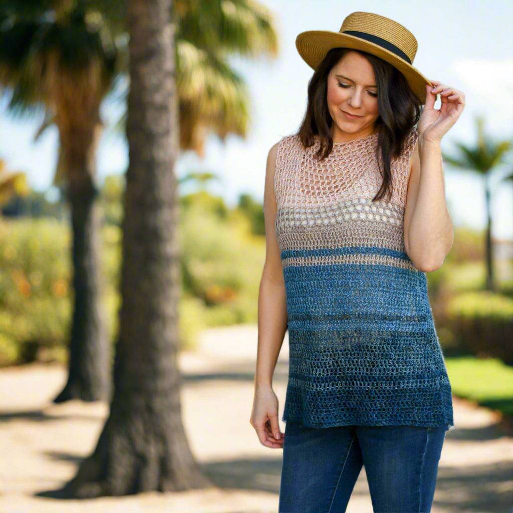 A woman wearing a sleeveless, relaxed-fit crochet top with an ombré gradient of pink, taupe, and navy blue against a beach backdrop.