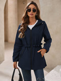A woman wearing a navy blue hooded waterproof windbreaker jacket with a gathered waist and zippered pockets.