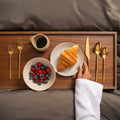 Emilia 20-piece flatware set in gold and silver finishes displayed on a wooden breakfast tray.