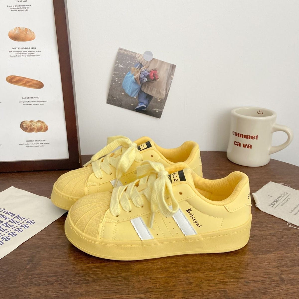 A pair of yellow shell toe sneakers with white stripes displayed on a wooden table, next to a coffee mug.