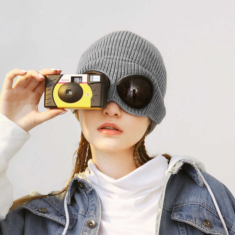 A woman wearing a grey ribbed knit pilot aviator hat with integrated goggle lenses, posing with a vintage camera.