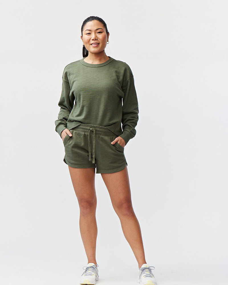 A woman wearing olive green rib-knit waistband shorts with pockets, standing in front of a white background.