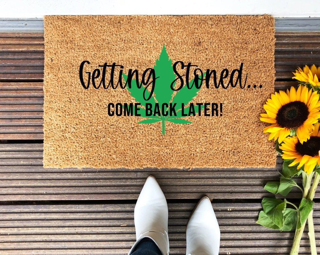 Natural coir doormat with 'Getting Stoned' text and a green leaf graphic sitting at a modern entryway.