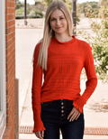 A woman wearing a vibrant sunset orange hollow-out knitted long sleeve top with ruffle detailing.