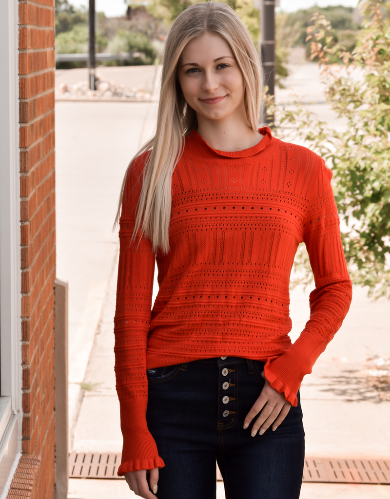A woman wearing a vibrant sunset orange hollow-out knitted long sleeve top with ruffle detailing.