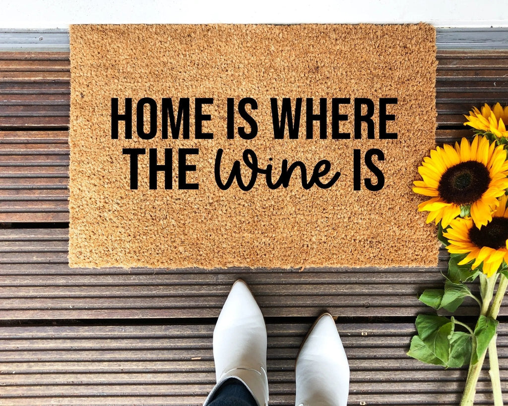 A natural coir doormat with the text Home Is Where The Wine Is positioned in a rustic entryway.