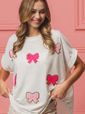 Close up of a woman wearing a white short-sleeve top with various sizes of bright pink embroidered bows featuring gold sequin trim.