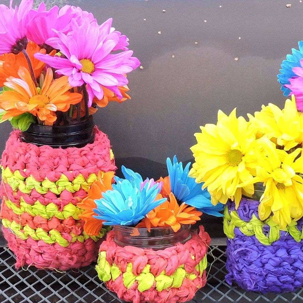 A crochet jar cozy wrapped around a glass mason jar on a wooden table.