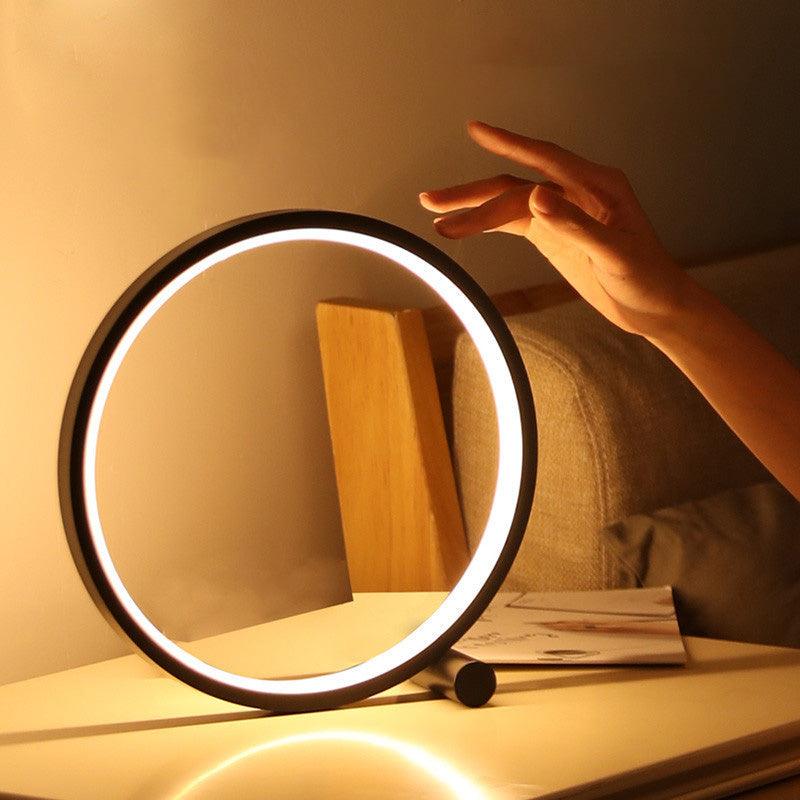 Minimalist round LED table lamp with a black and white iron base and acrylic shade on a nightstand.