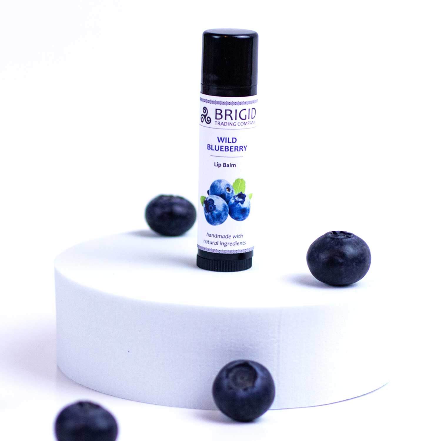 A tube of Wild Blueberry lip balm surrounded by fresh blueberries on a white pedestal.