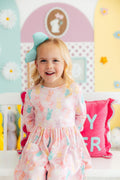A smiling young girl wearing a pink 3/4 sleeve dress with a bunny and flower print, sitting on a white bench.