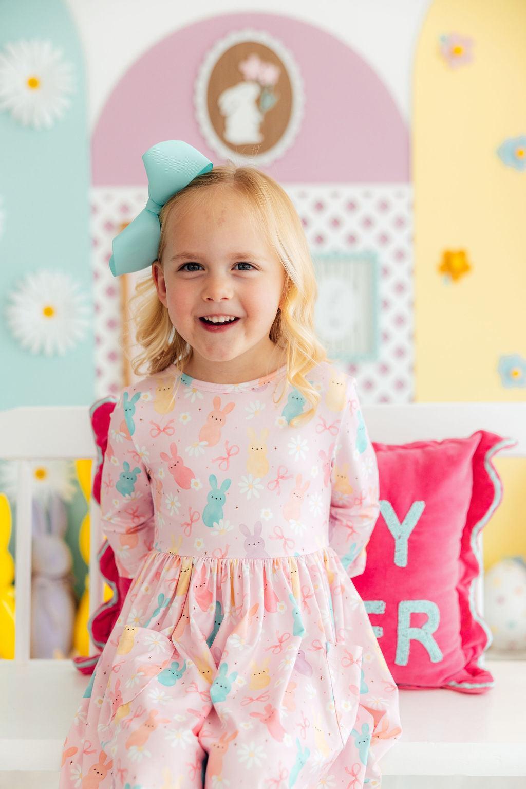 A smiling young girl wearing a pink 3/4 sleeve dress with a bunny and flower print, sitting on a white bench.