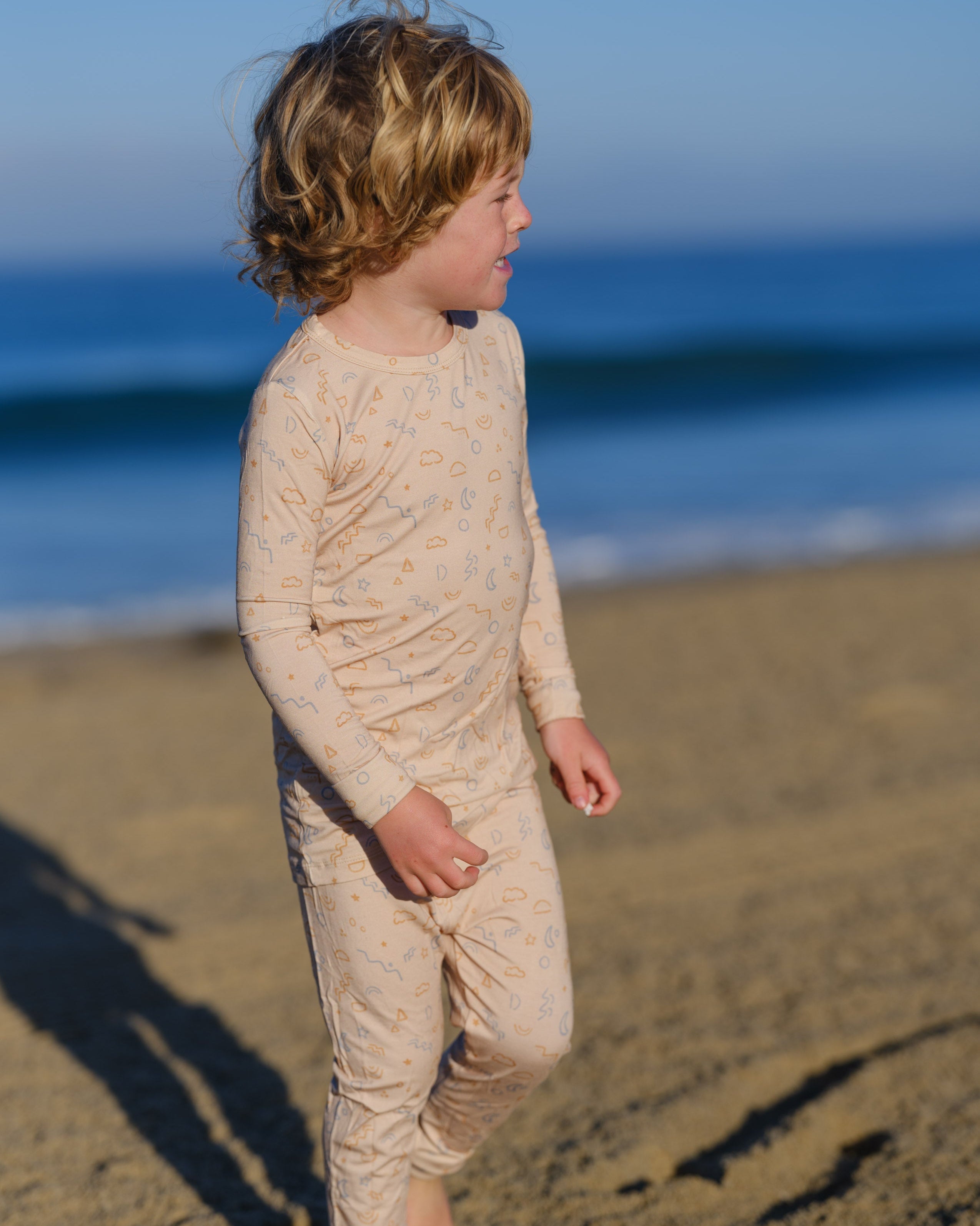 Child wearing a beige long-sleeve shirt and pants standing on a beach with ocean in the background