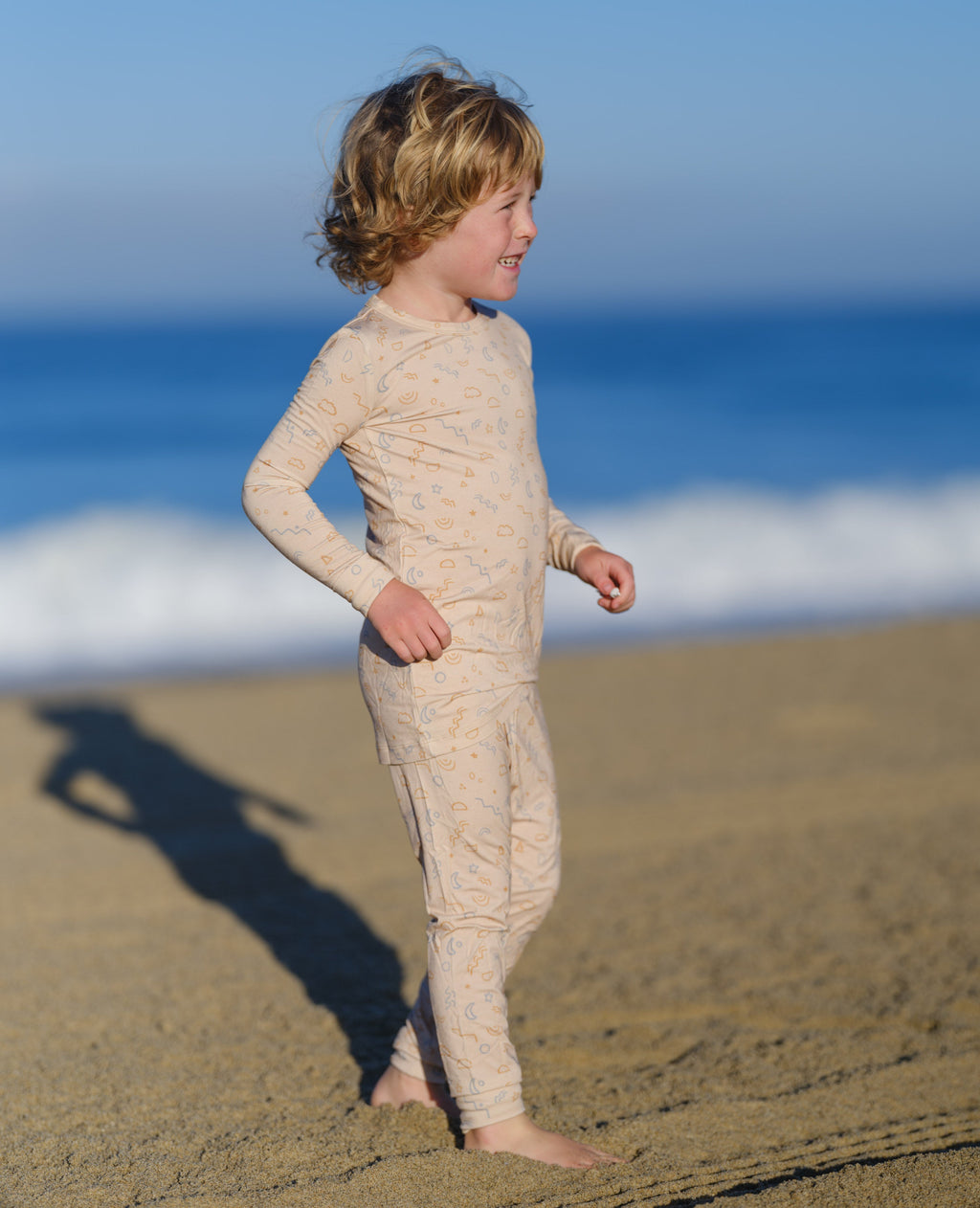 Child wearing a beige long-sleeve shirt and pants on a sandy beach with ocean waves in the background.