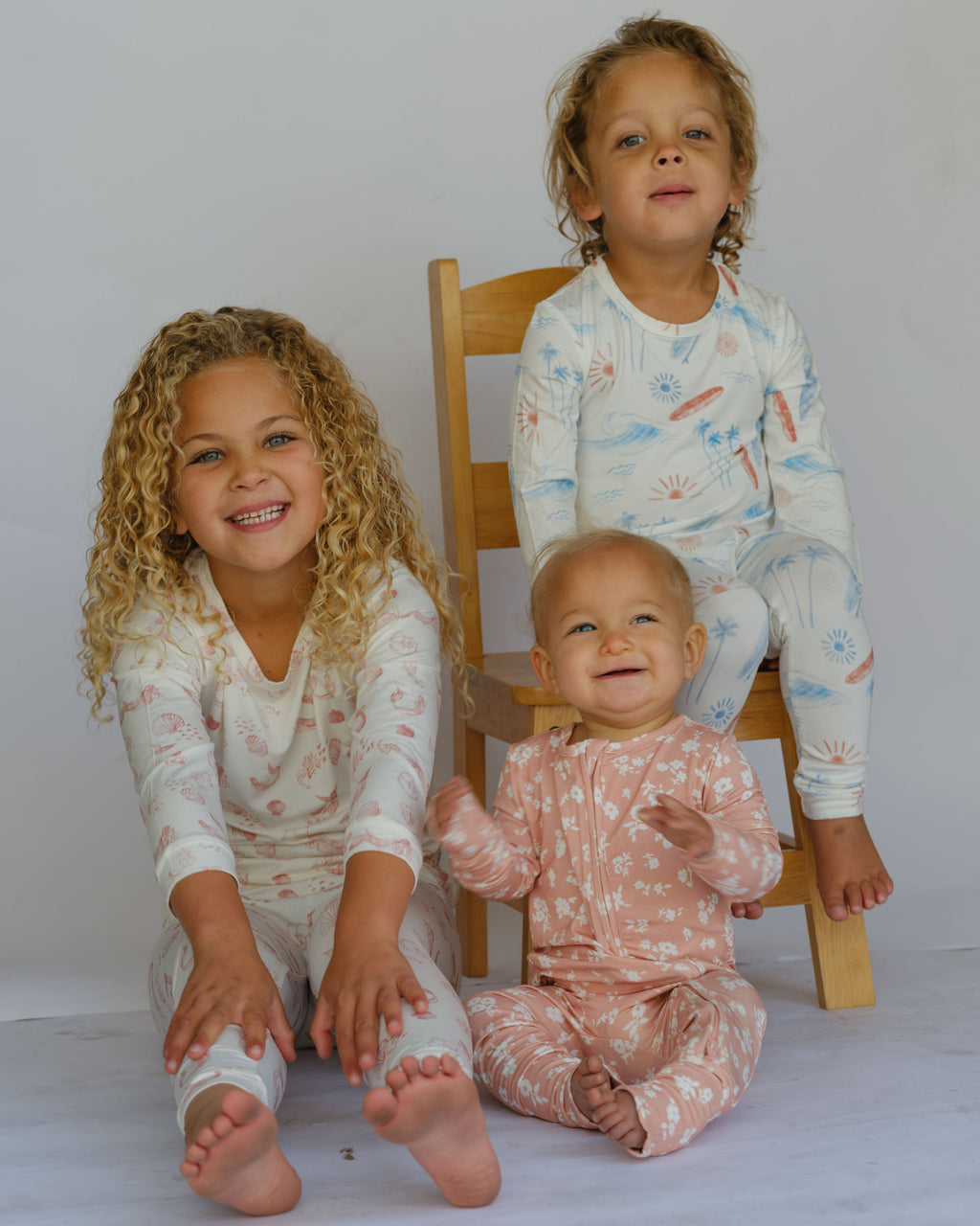 Three children in matching pajamas sitting on a white floor with a wooden chair.