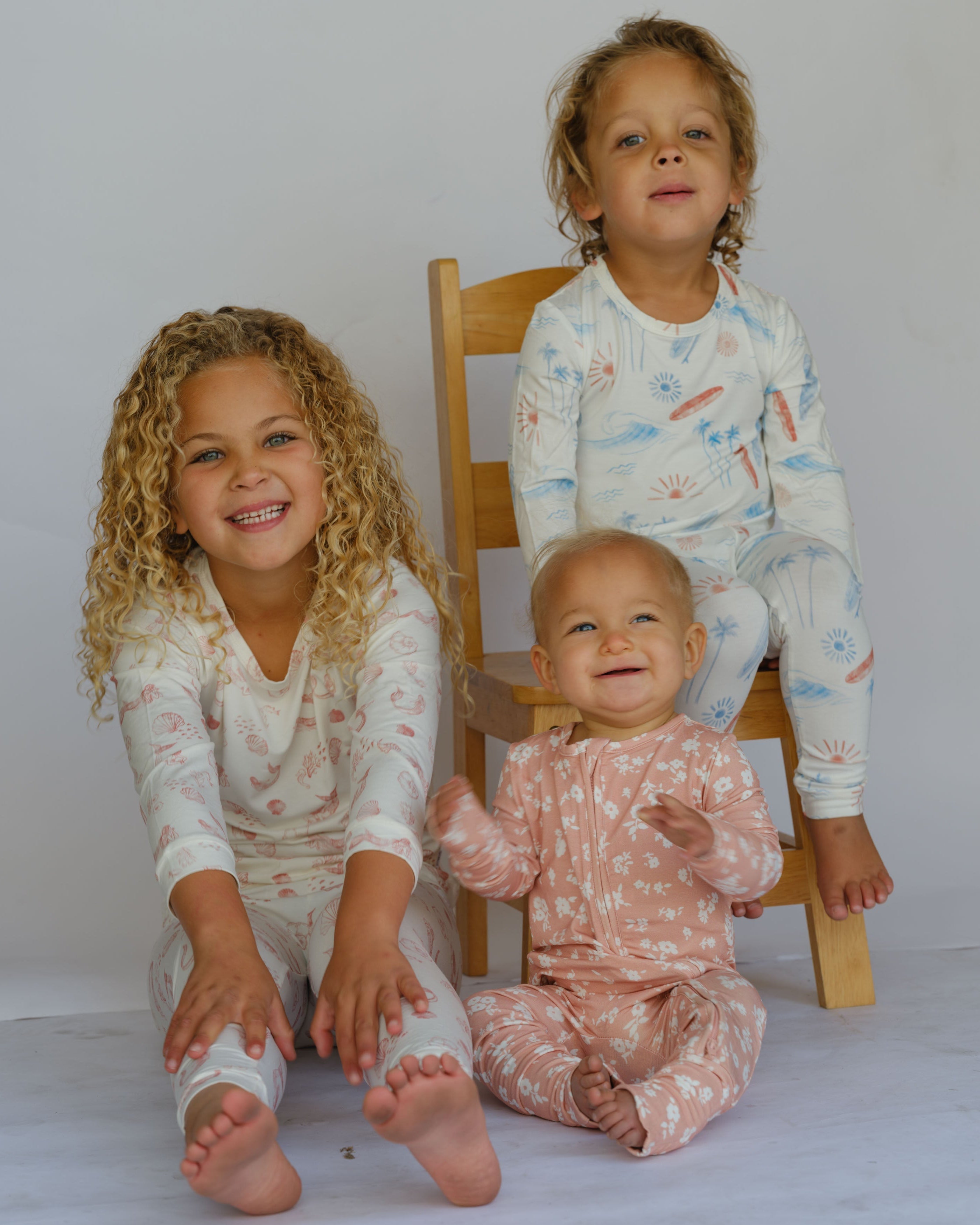 Three children in matching pajamas sitting on a white floor with a wooden chair.