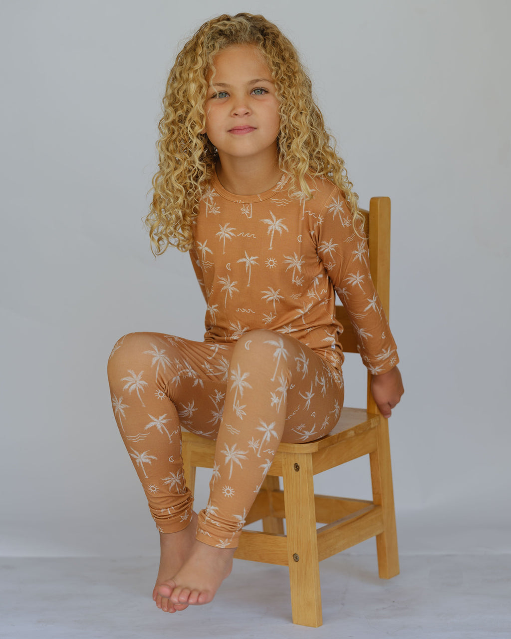 Child wearing a brown patterned outfit sitting on a wooden chair against a plain background