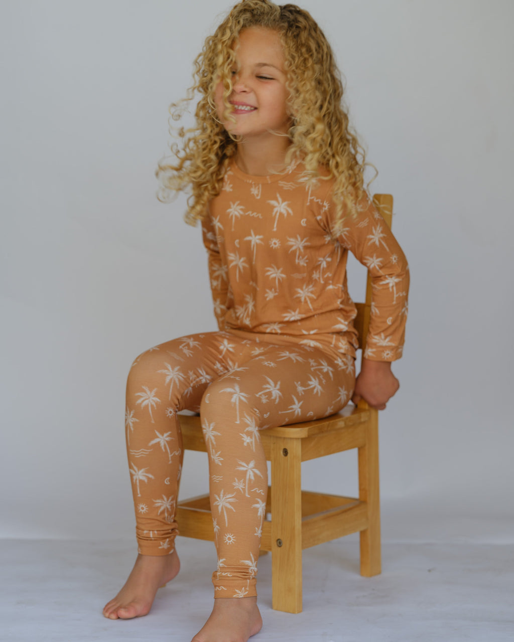 Child wearing a brown pajama set with white floral patterns, sitting on a wooden stool against a plain background.