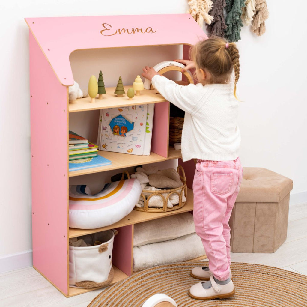Pink and white Montessori-inspired dollhouse bookcase featuring open shelving in a child's bedroom.