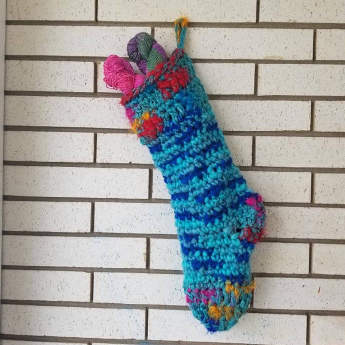 Vibrant handmade Punky Chunky crochet holiday stocking hanging on a mantle, showcasing blue, pink, yellow, and red abstract patterns.
