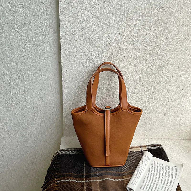 Brown bucket style shoulder bag with a metallic lock detail sitting on a table.