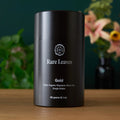A cylindrical black tea tin and a green tea box, both labeled Rare Leaves Gold, sitting on a wooden surface.