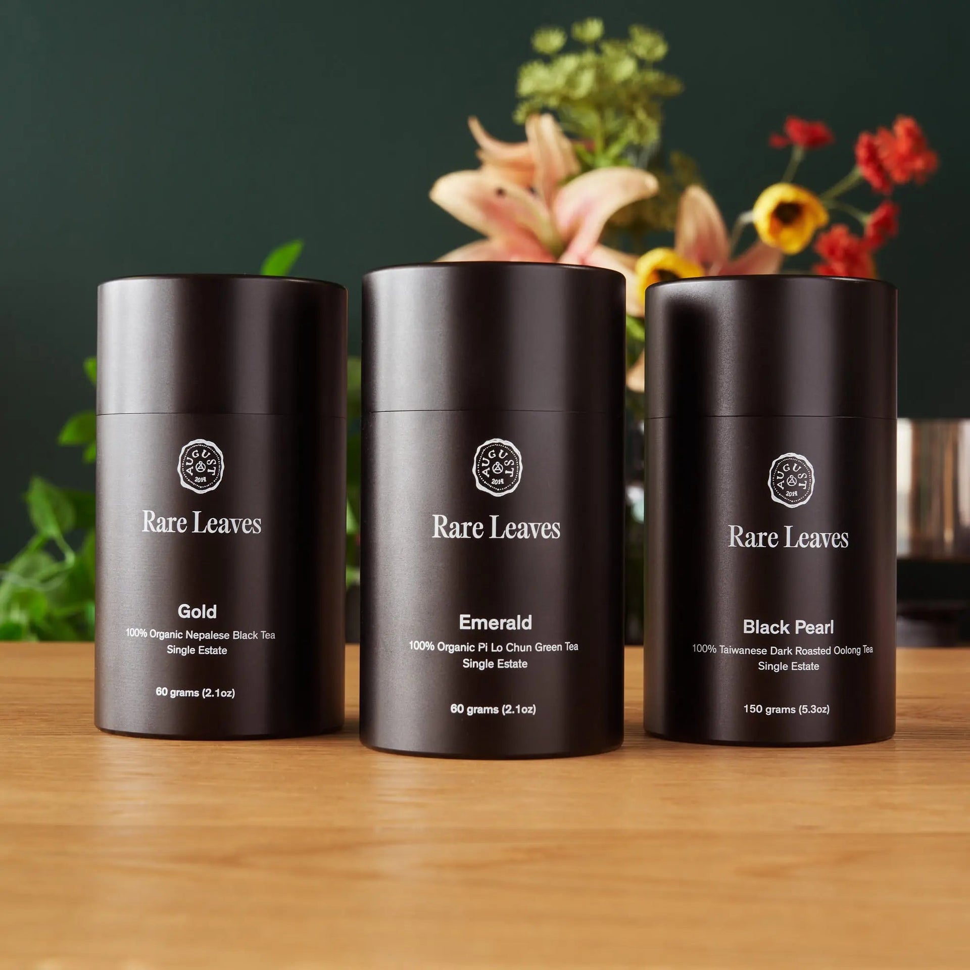 Three dark canisters of Rare Leaves single origin tea displayed on a wooden table with floral accents