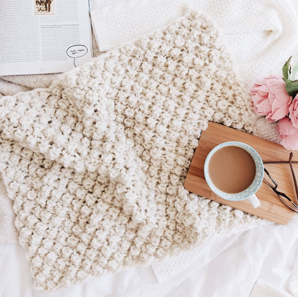 A soft, cream-colored Raspberry Dreams knit baby blanket styled in a modern, minimalist nursery setting.