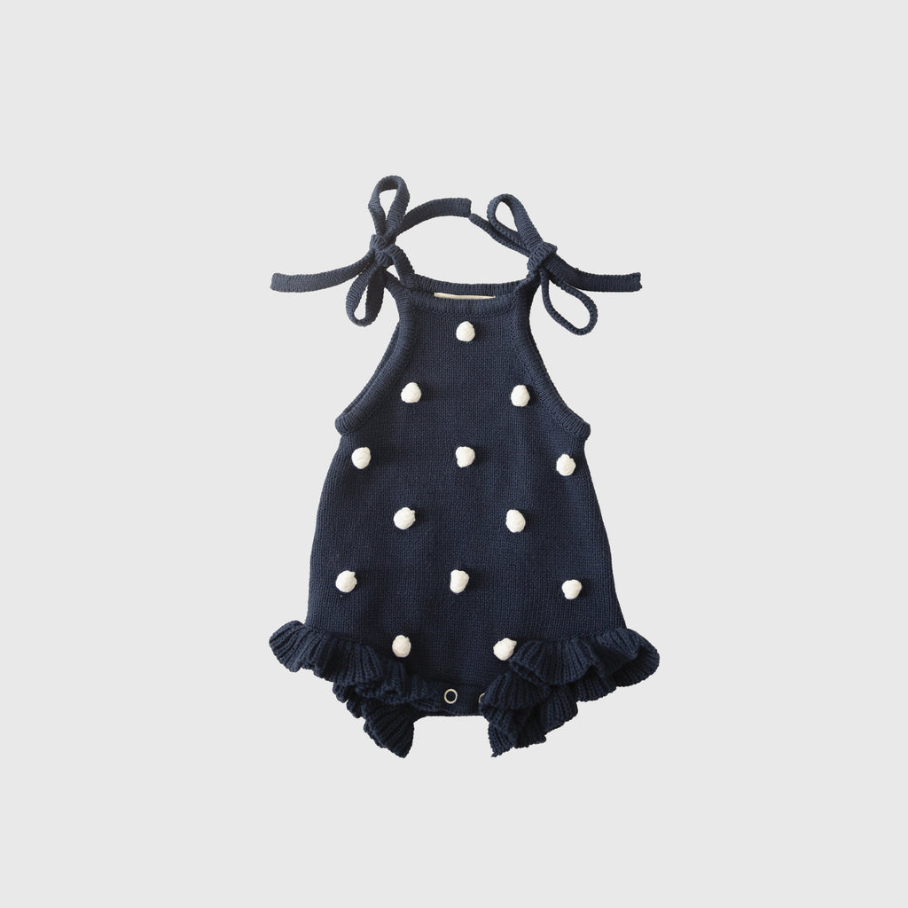 Navy blue knit baby romper with cream pom-poms, ruffle details, and tie straps