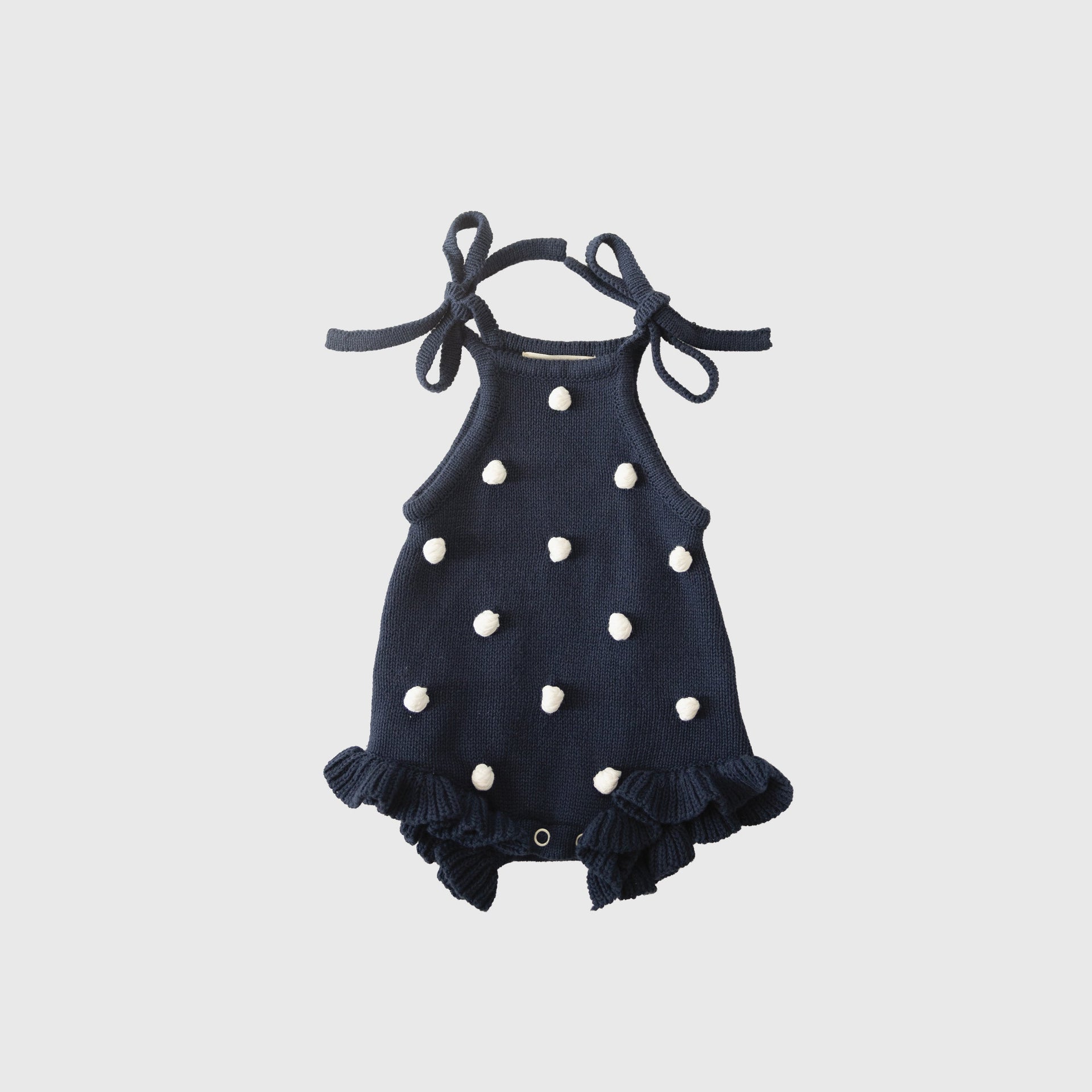 Navy blue knit baby romper with cream pom-poms, ruffle details, and tie straps