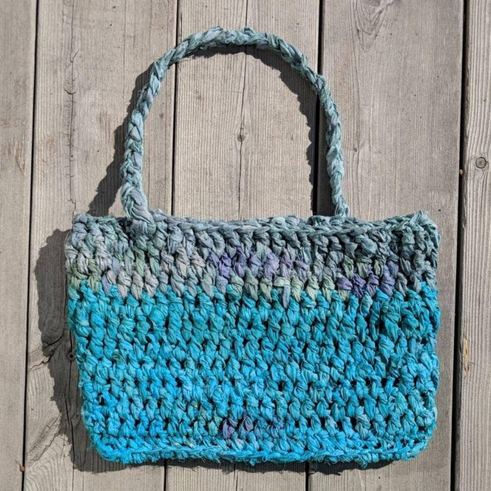 A handmade bohemian beach bag in teal, turquoise, and blue-grey sitting on a sandy surface.