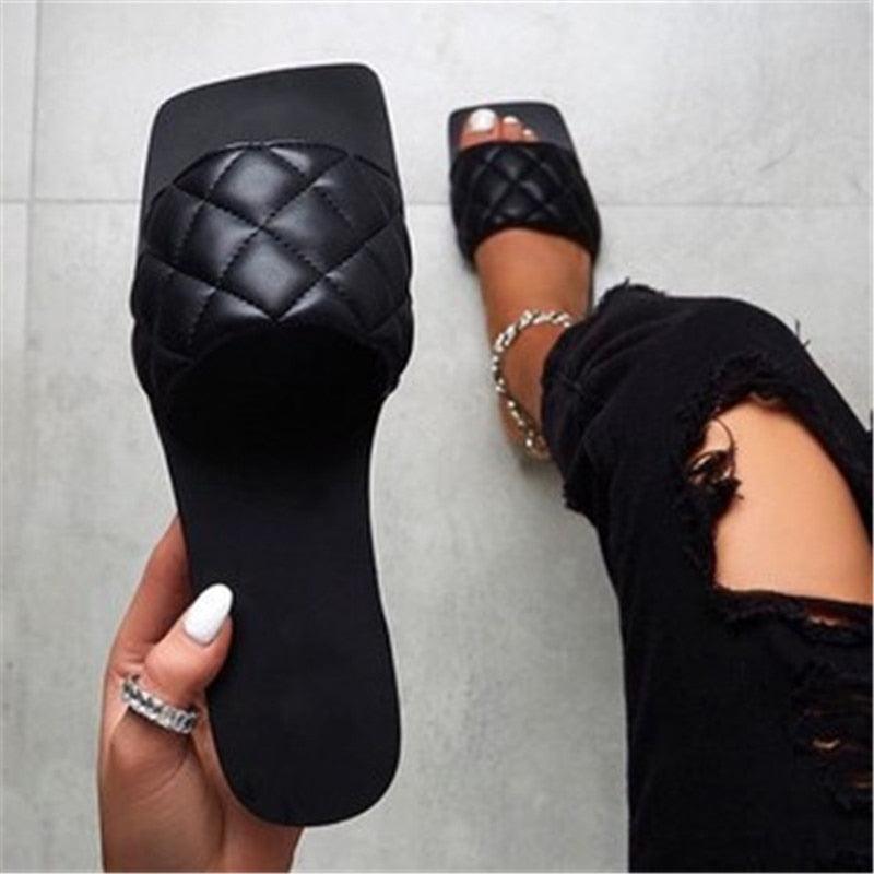 A pair of minimalist woven leather flat slides for women in beige, black, and metallic-gold, captured in a lifestyle resort setting.