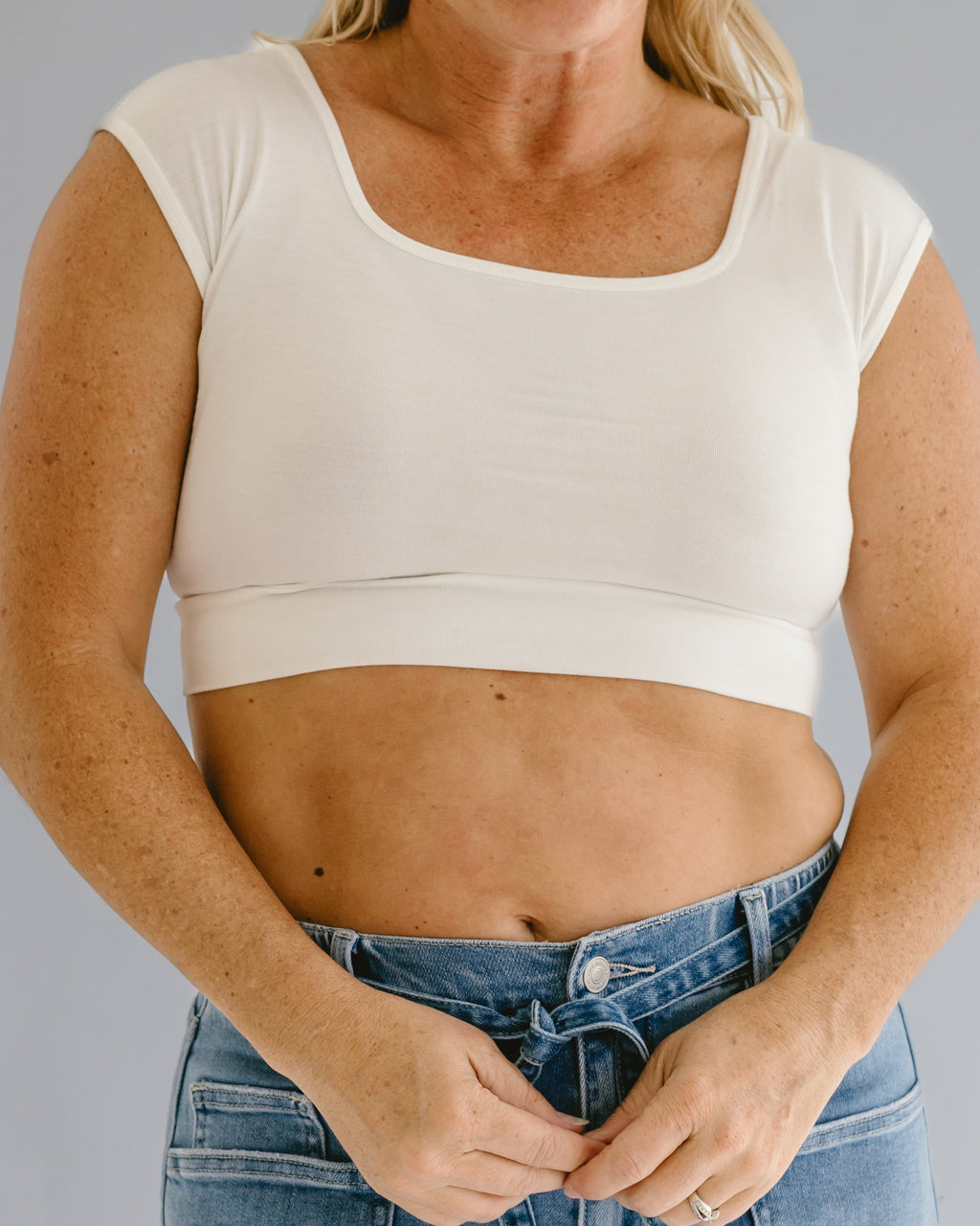A woman wearing a white square neck cap sleeve layering top underneath a pair of jeans.