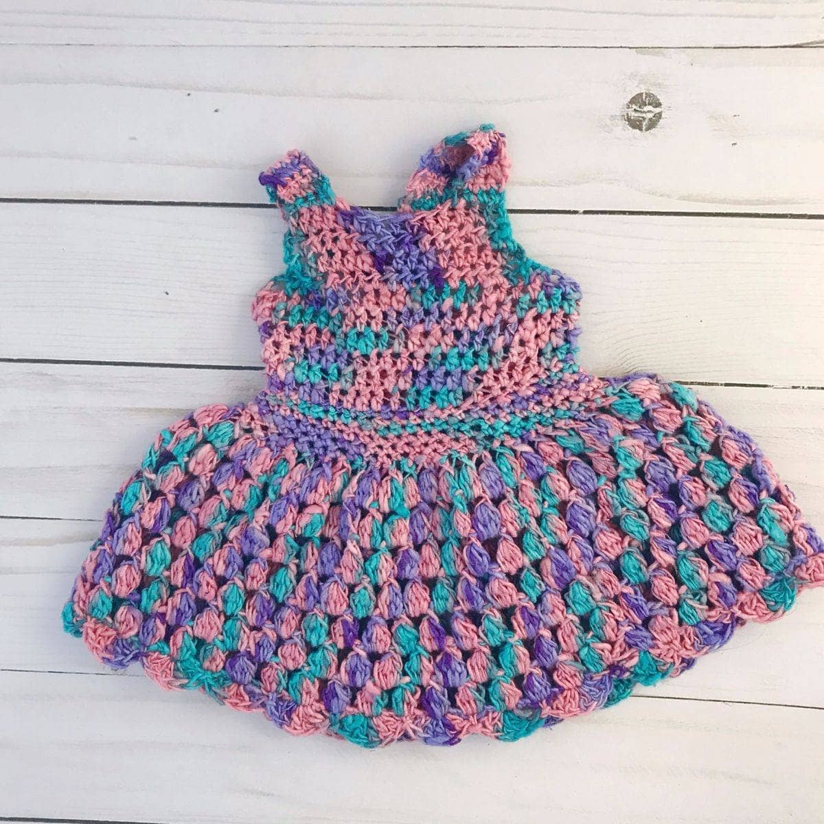 A handmade crochet sundress for an 18-inch doll in variegated pink, purple, and turquoise yarn with a scoop neck and button closure.