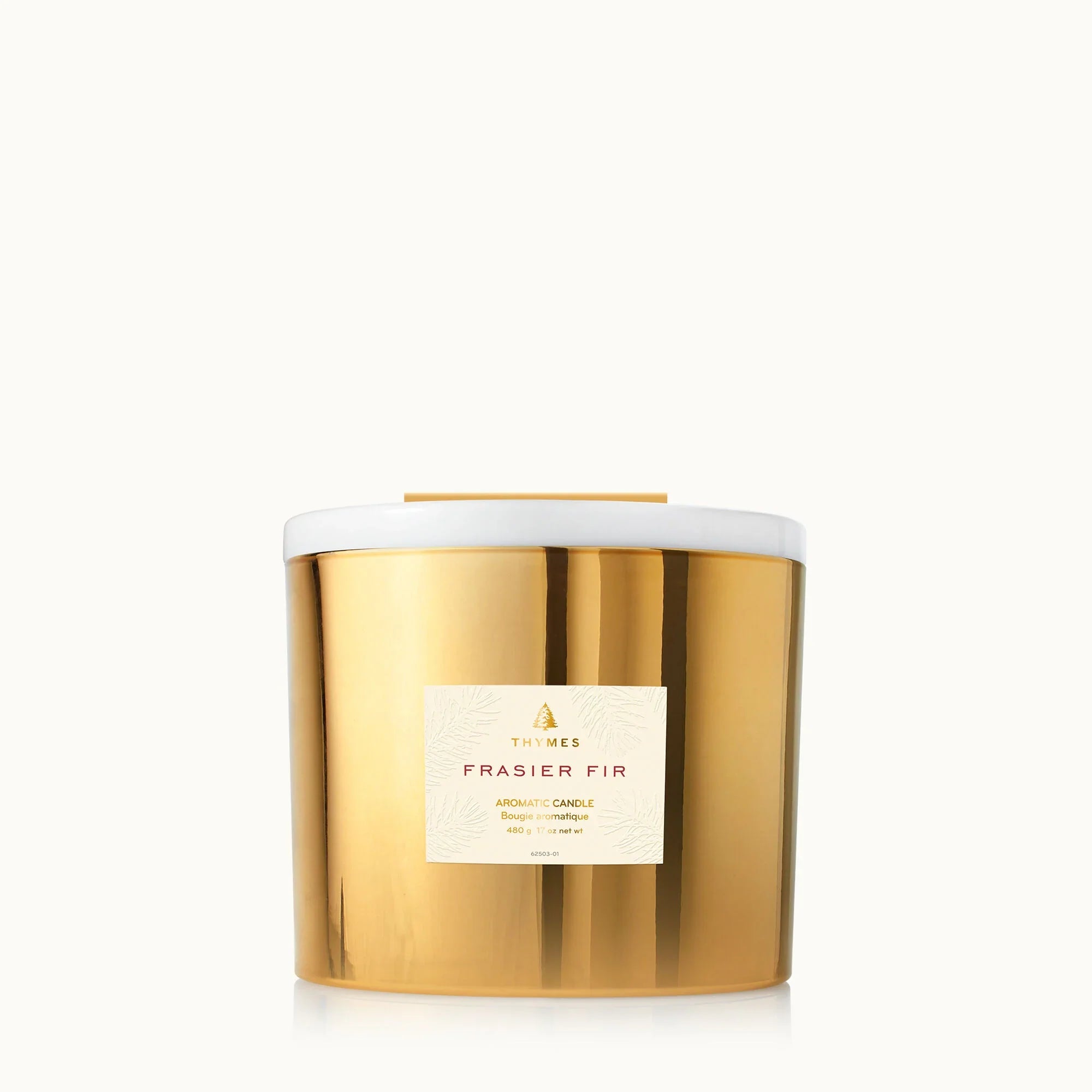 A gold 3-wick Frasier Fir candle sitting on a wooden surface with seasonal decor.