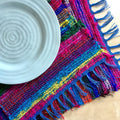 Colorful bohemian-style woven placemat with vibrant striped pattern in hot pink, electric blue, lime green, yellow, and orange.