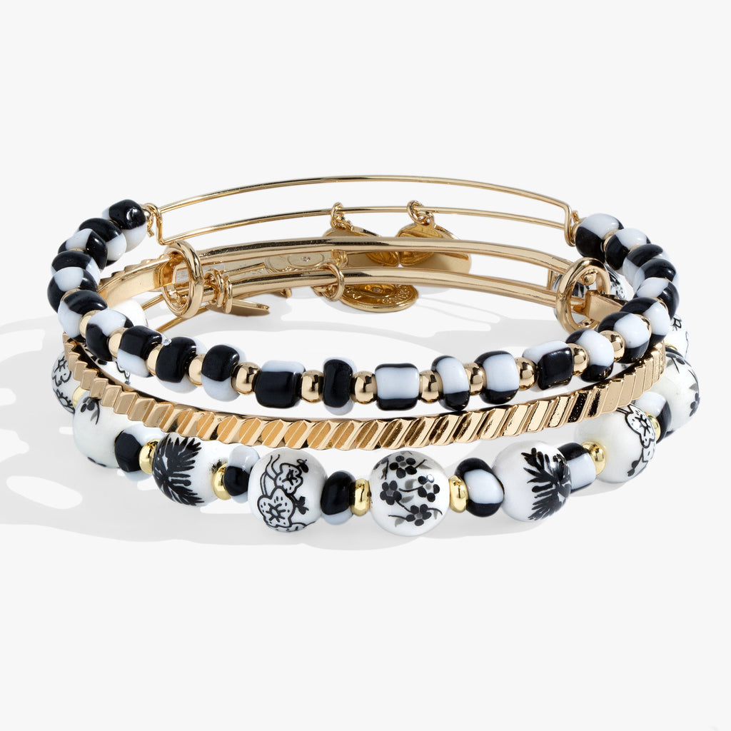 Set of three bangles featuring one hand-painted toile floral pattern, one black and white glass beaded bangle, and one gold-tone textured bangle.