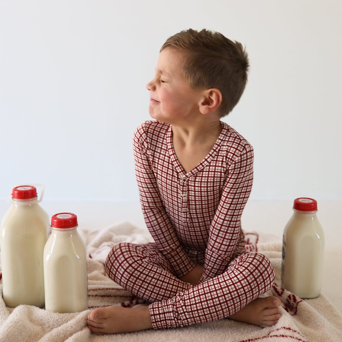 A young boy wearing a red and white window pane patterned two-piece bamboo pajama set while sitting on a blanket.